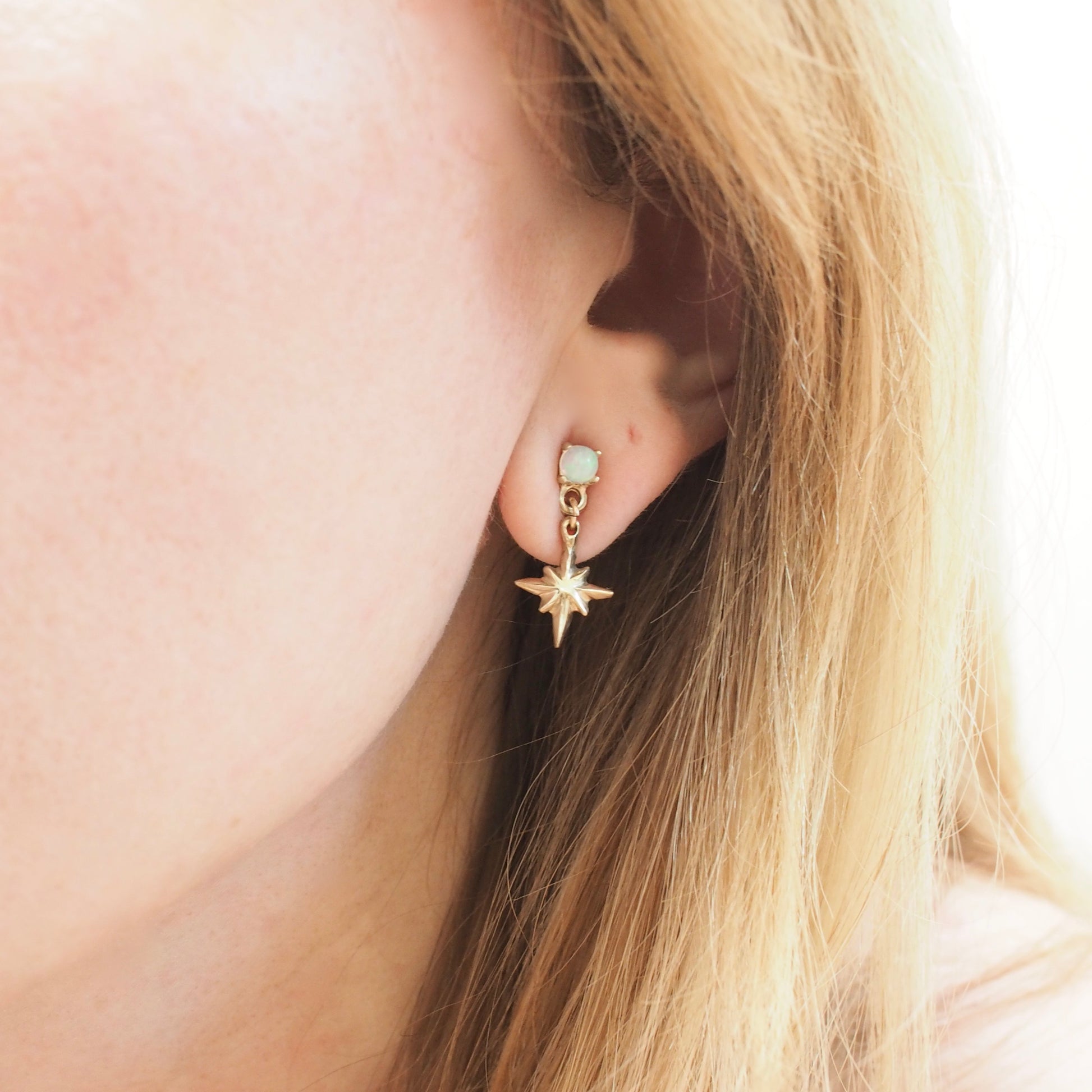 Tiny dangly star earrings set with 4mm sustainably sourced opals, in Gold Tone bronze. Made by Iron Oxide Designs and shown on a model for scale