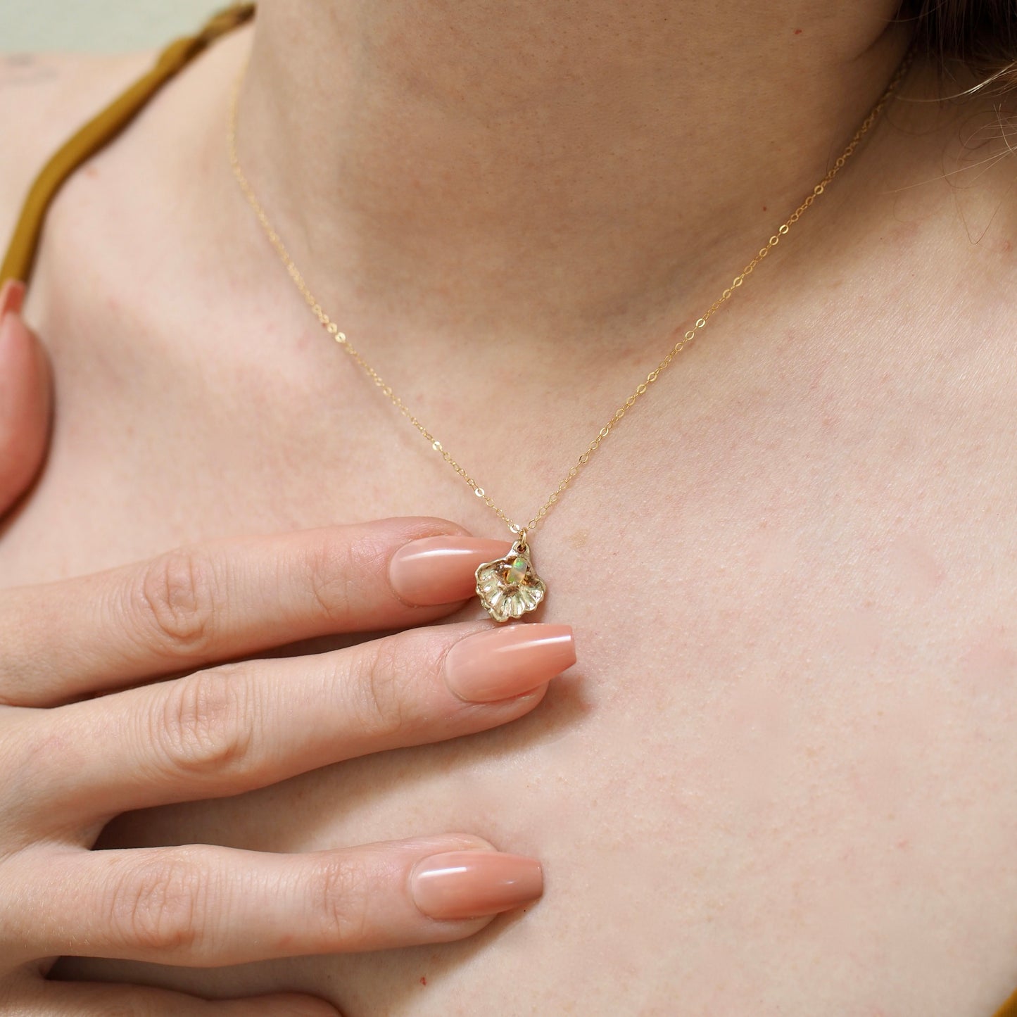 Close up of a gold tone Seashell necklace containing a tiny opal in place of a pearl, handmade by Iron Oxide Designs, shown on a. model for scale