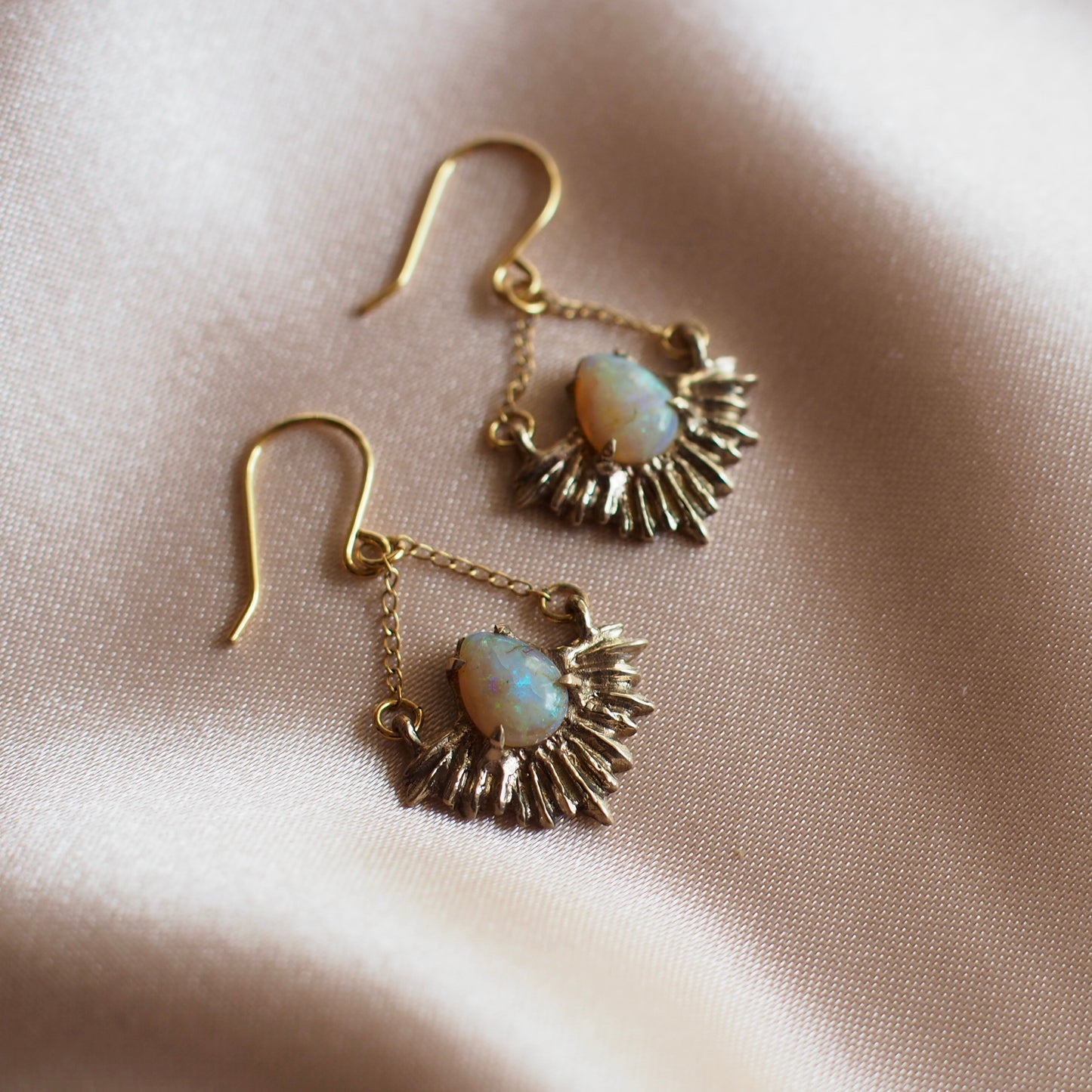 Gold Tone bronze earrings in the shape of beams of light set with teardrop shaped, lab grown opals on a pink satin background