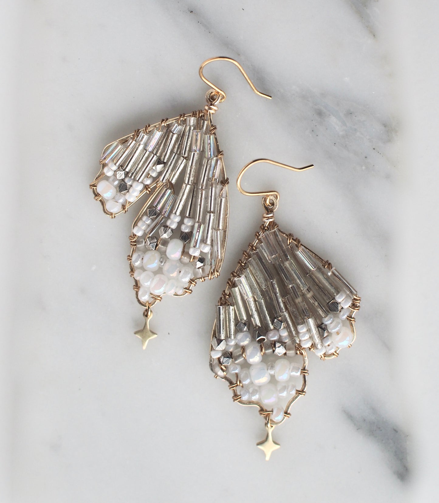 A close-up view of handmade celestial earring featuring a delicate wing-shaped design. The earring is crafted with shimmering metallic beads, opalescent accents, and a small dangling star charm, hanging elegantly from a gold hook.