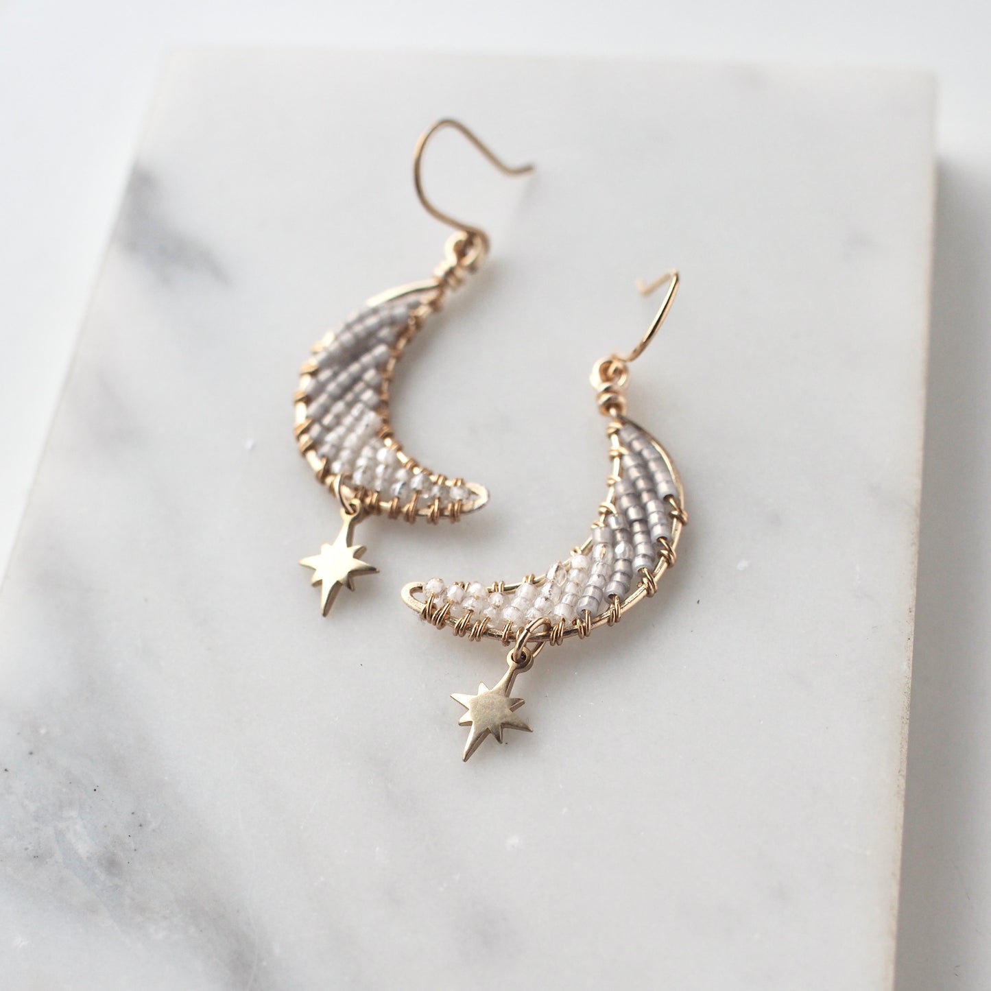 A close-up view of handmade celestial earring featuring a delicate moon-shaped design on a marble tray. The elegant earrings are crafted with bronze star charms, and tiny toho beads. The color of the beads ranges from white to dusky gray.