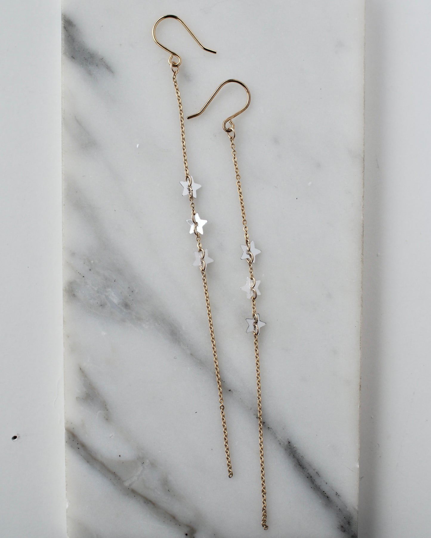 A close-up view of handmade dangle  earrings on a slab of marble. These ethereal earrings featuring three star shaped sequins set on a fine, 5" gold filled chain.