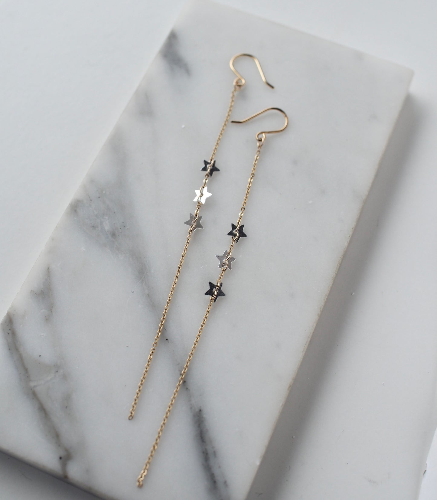 A close-up view of handmade dangle  earrings on a slab of marble. These ethereal earrings featuring three star shaped sequins set on a fine, 5" gold filled chain.