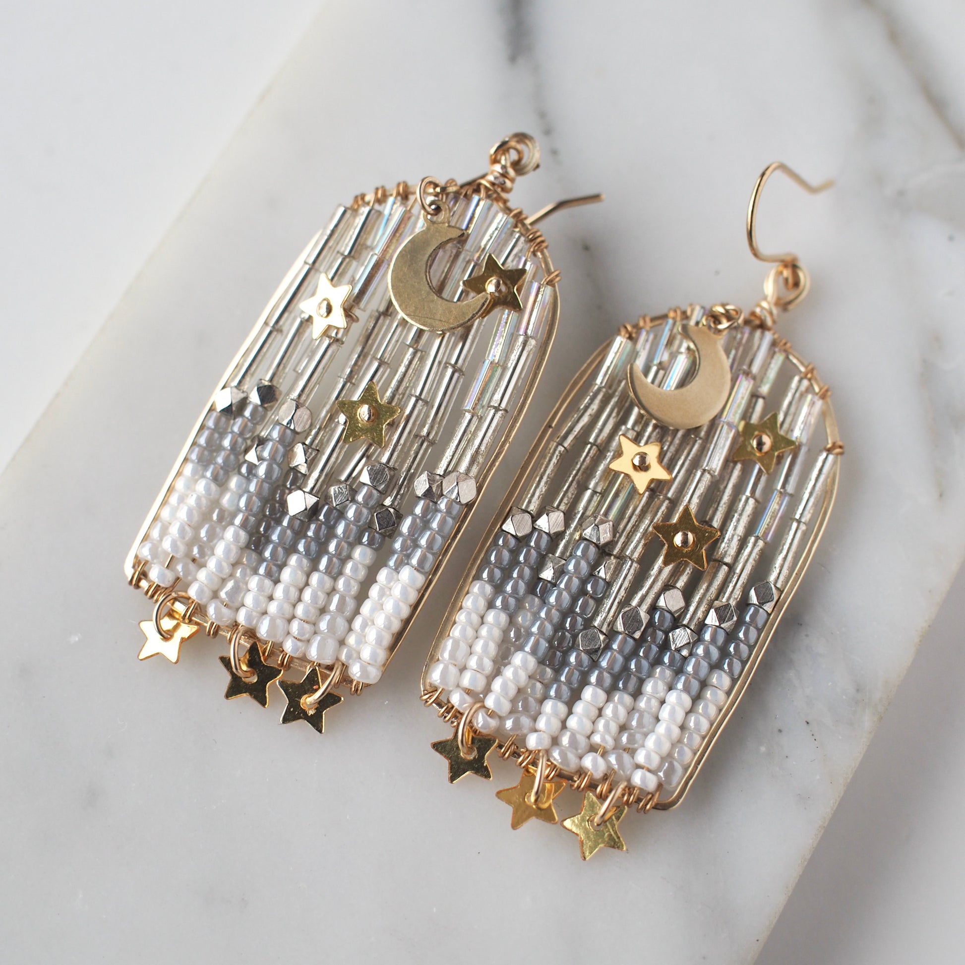 A close-up view of handmade celestial earrings featuring an archway shaped design with a moon and stars on each earring. The ethereal portal earrings are crafted with shimmering metallic star sequins, opalescent beads, and small dangling stars.