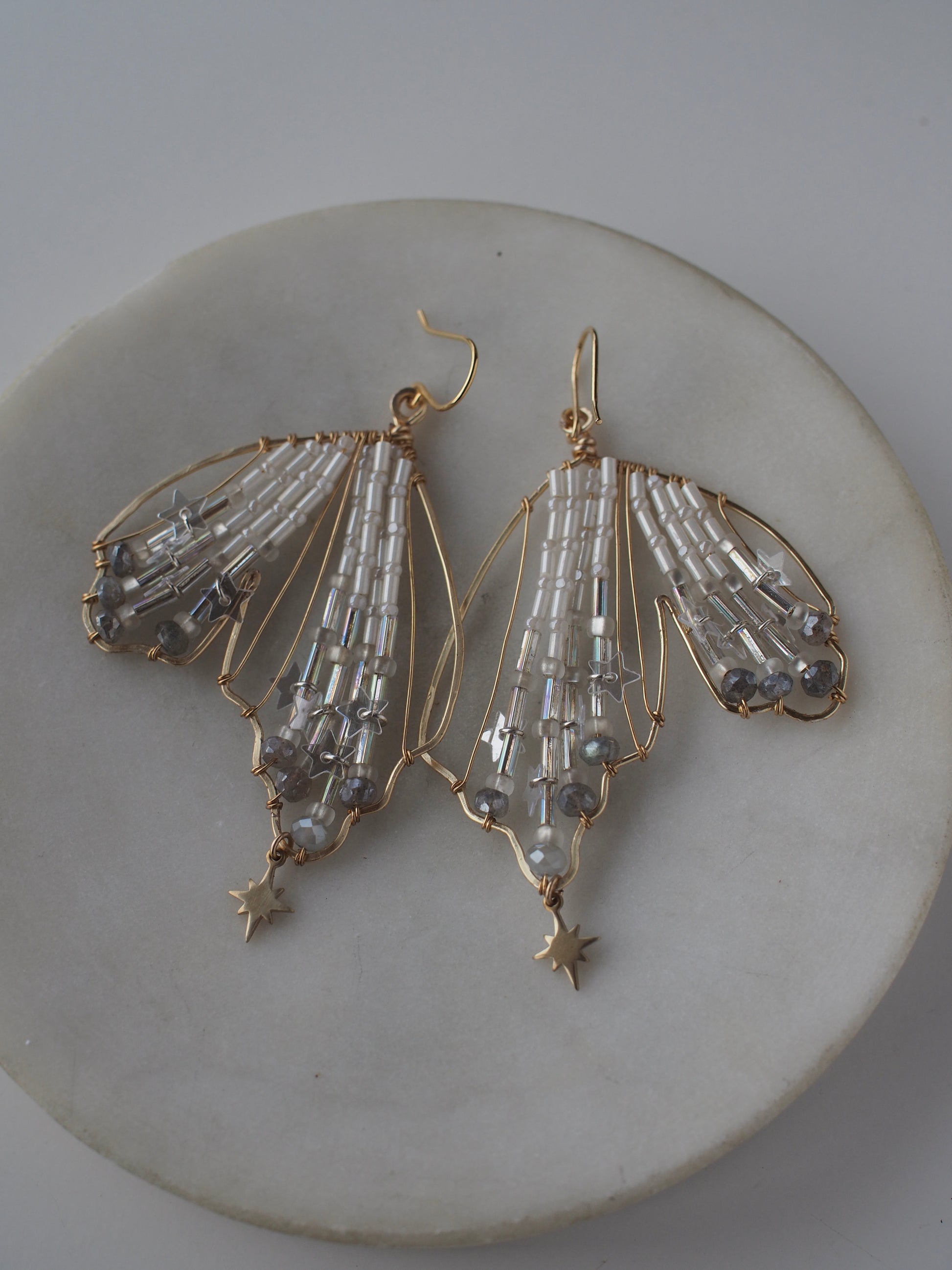 A close-up view of handmade celestial earring featuring a delicate wing-shaped design. The ethereal earrings are crafted with shimmering metallic star sequins, opalescent beads, and small dangling stars.