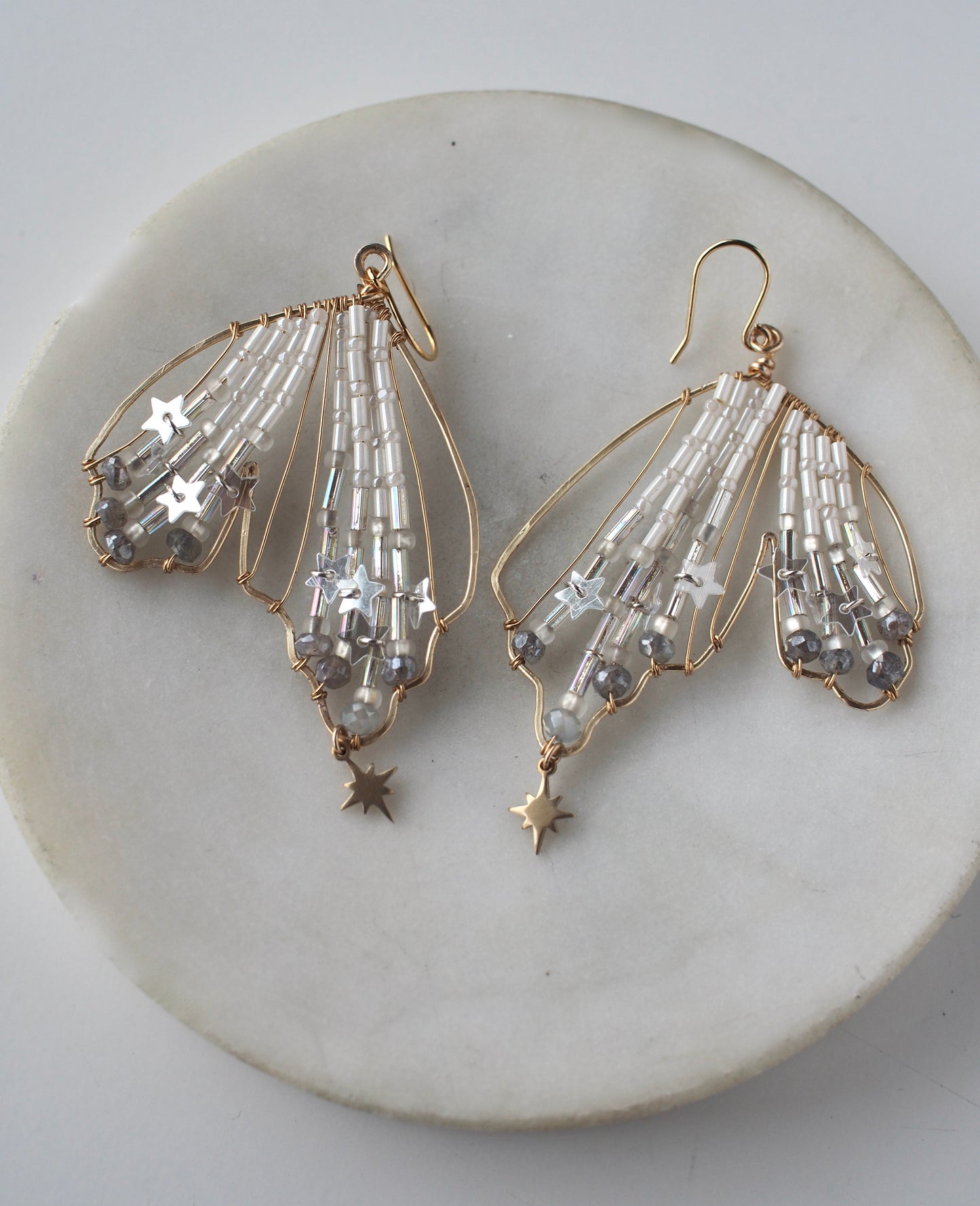 A close-up view of handmade celestial earring featuring a delicate wing-shaped design on a round marble dish. The elegant earrings are crafted with shimmering metallic star sequins, opalescent beads, and small dangling stars.