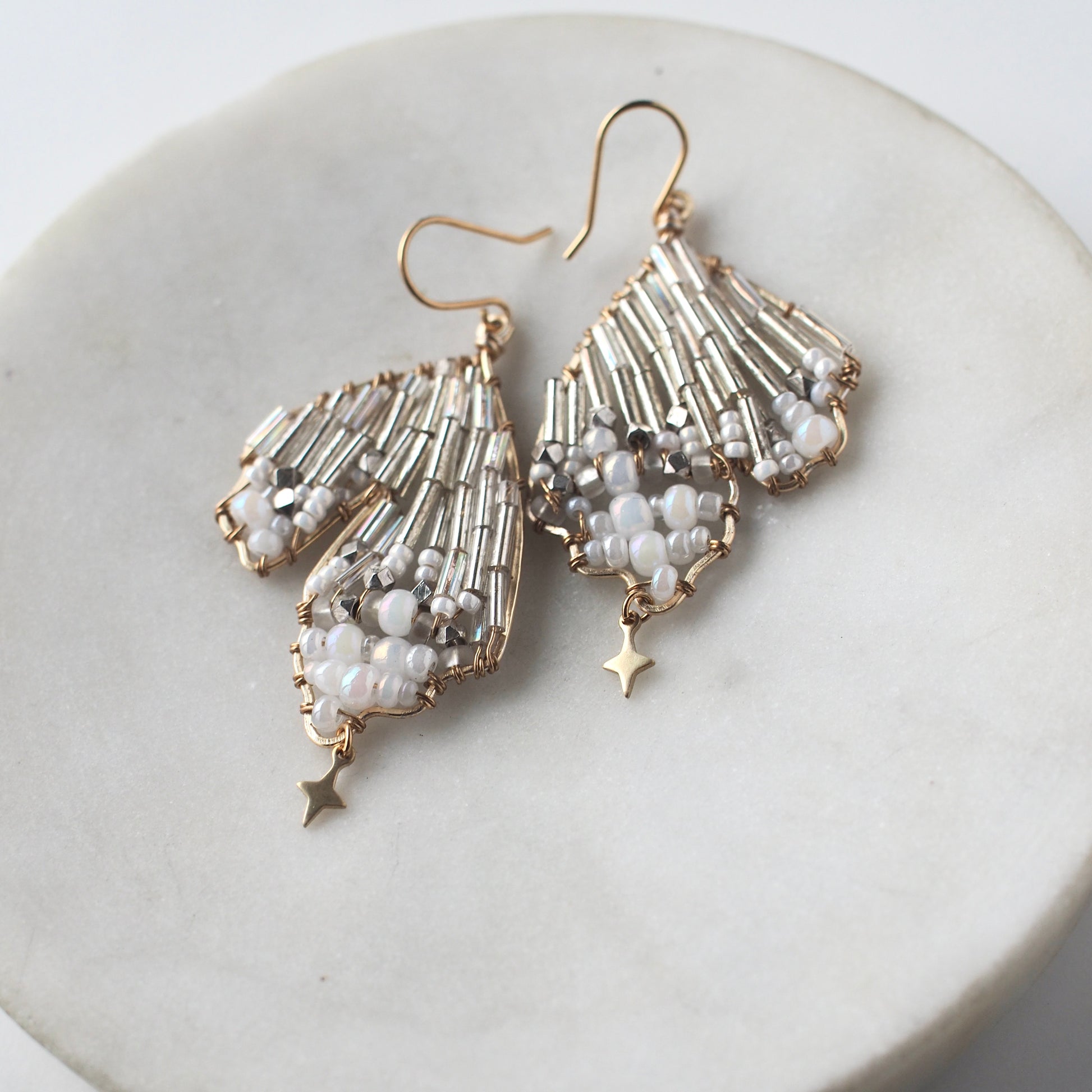 A close-up view of handmade celestial earring featuring a delicate wing-shaped design on a round marble dish. The earrings are crafted with shimmering metallic beads, opalescent accents, and small dangling stars, hanging elegantly from a gold hook.