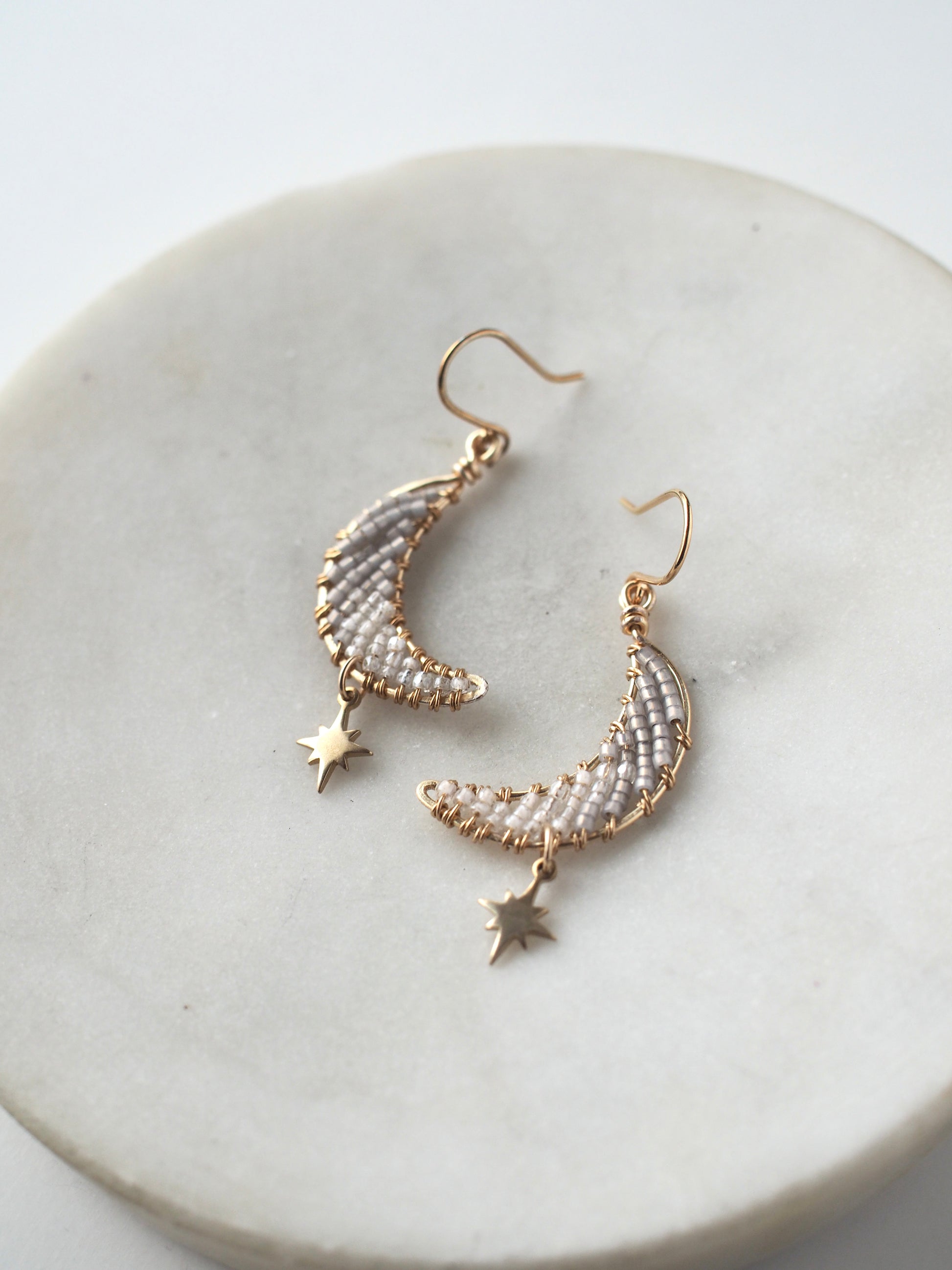 A close-up view of handmade celestial earring featuring a delicate moon-shaped design on a round marble tray. The elegant earrings are crafted with bronze star charms, and tiny toho beads. The color of the beads ranges from white to dusky gray.