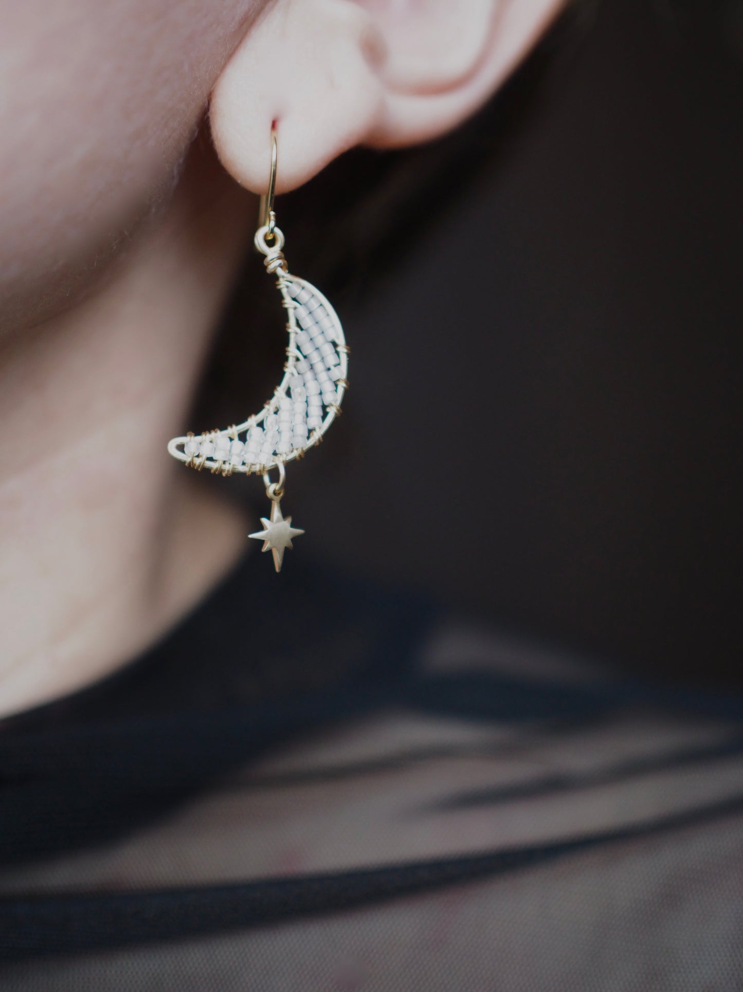 A close-up of someone wearing handmade celestial earring featuring a delicate moon-shaped design on a marble tray. The elegant earrings are crafted with bronze star charms, and tiny toho beads. The color of the beads ranges from white to dusky gray.