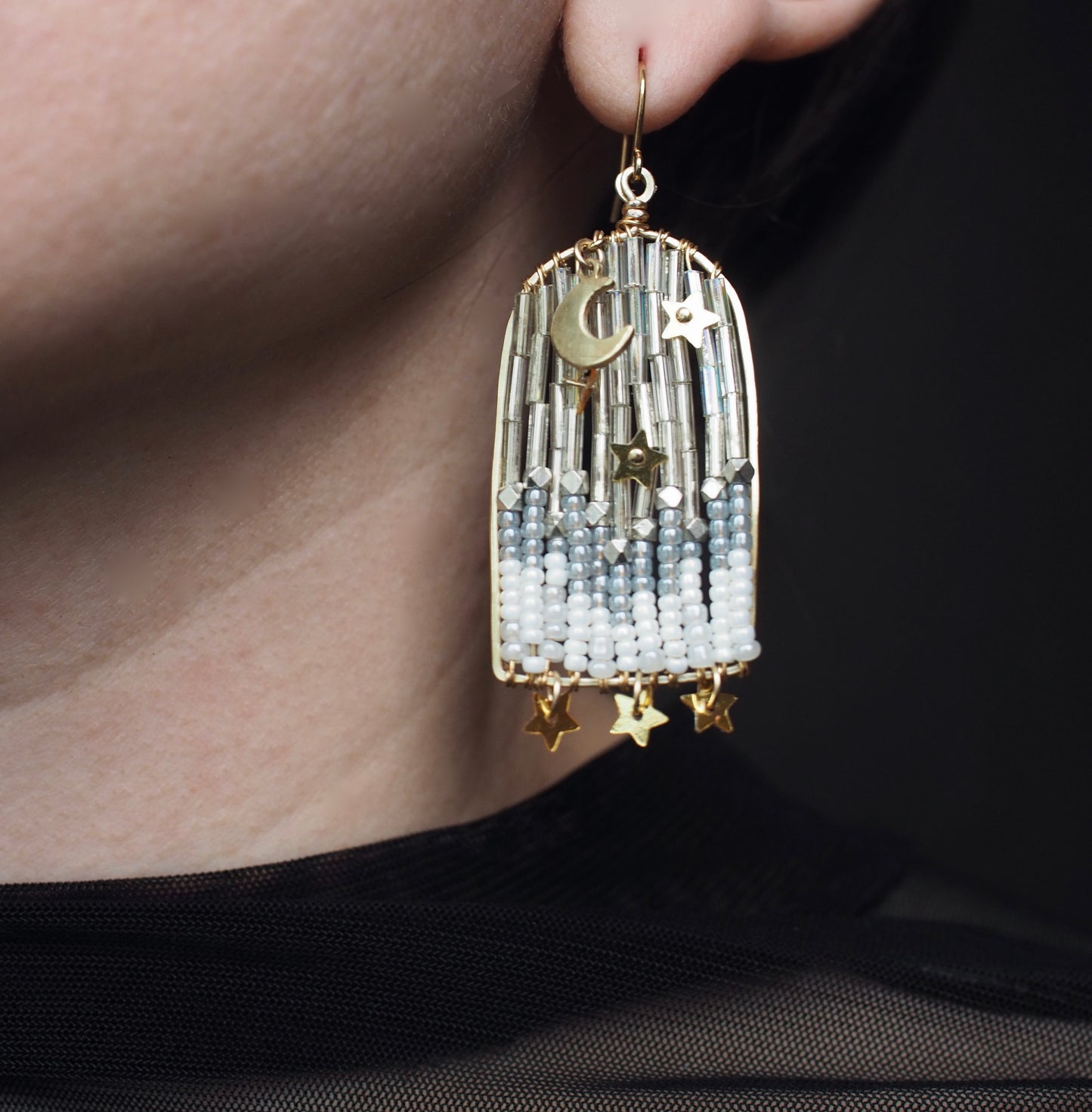 A close-up view of someone wearing handmade celestial earrings featuring an archway shaped design with a moon and stars. The ethereal portal earrings are crafted with shimmering metallic star sequins, opalescent beads, and small dangling stars.