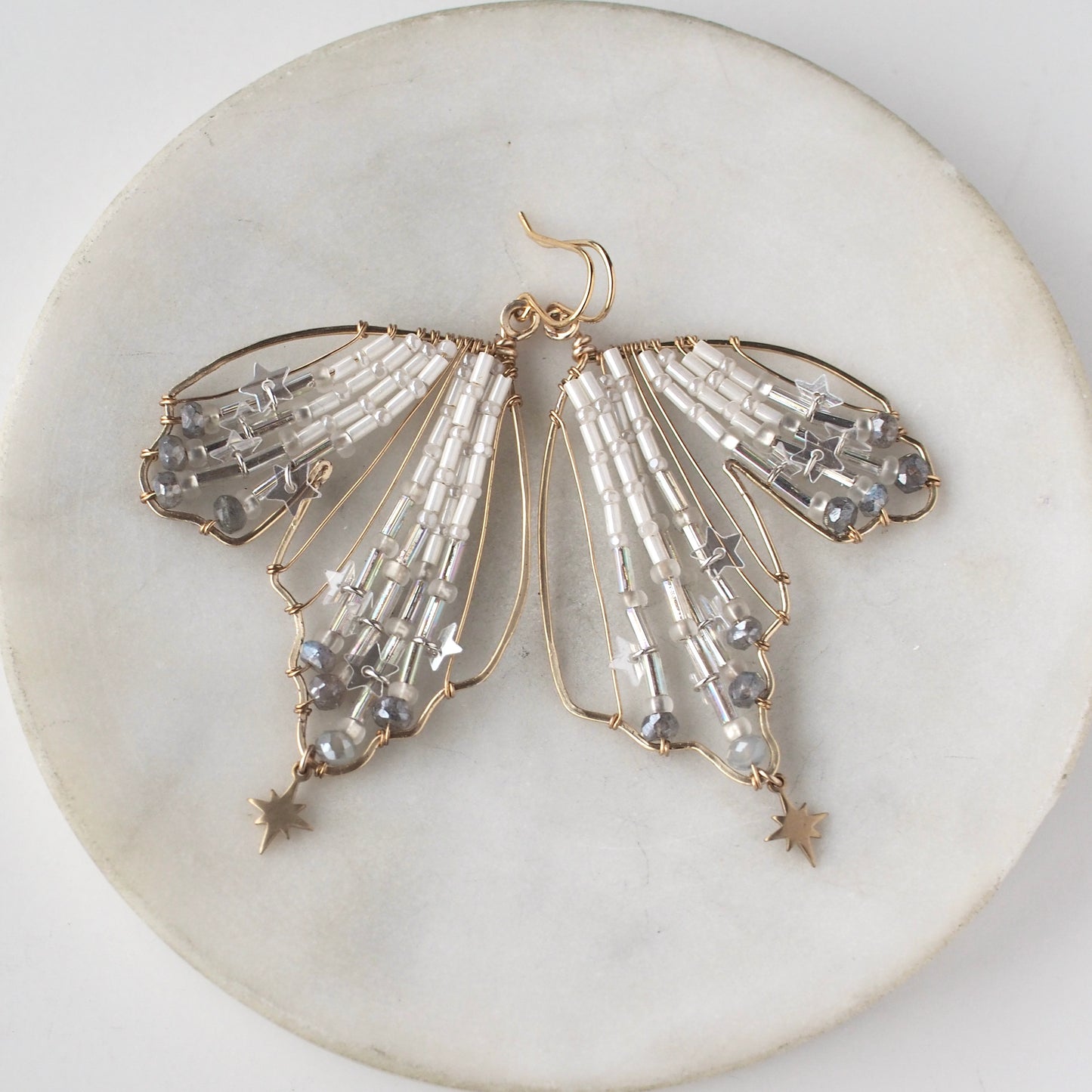 A close-up view of handmade celestial earring featuring a delicate wing-shaped design on a round marble dish. The elegant earrings are crafted with shimmering metallic star sequins, opalescent beads, and small dangling stars.