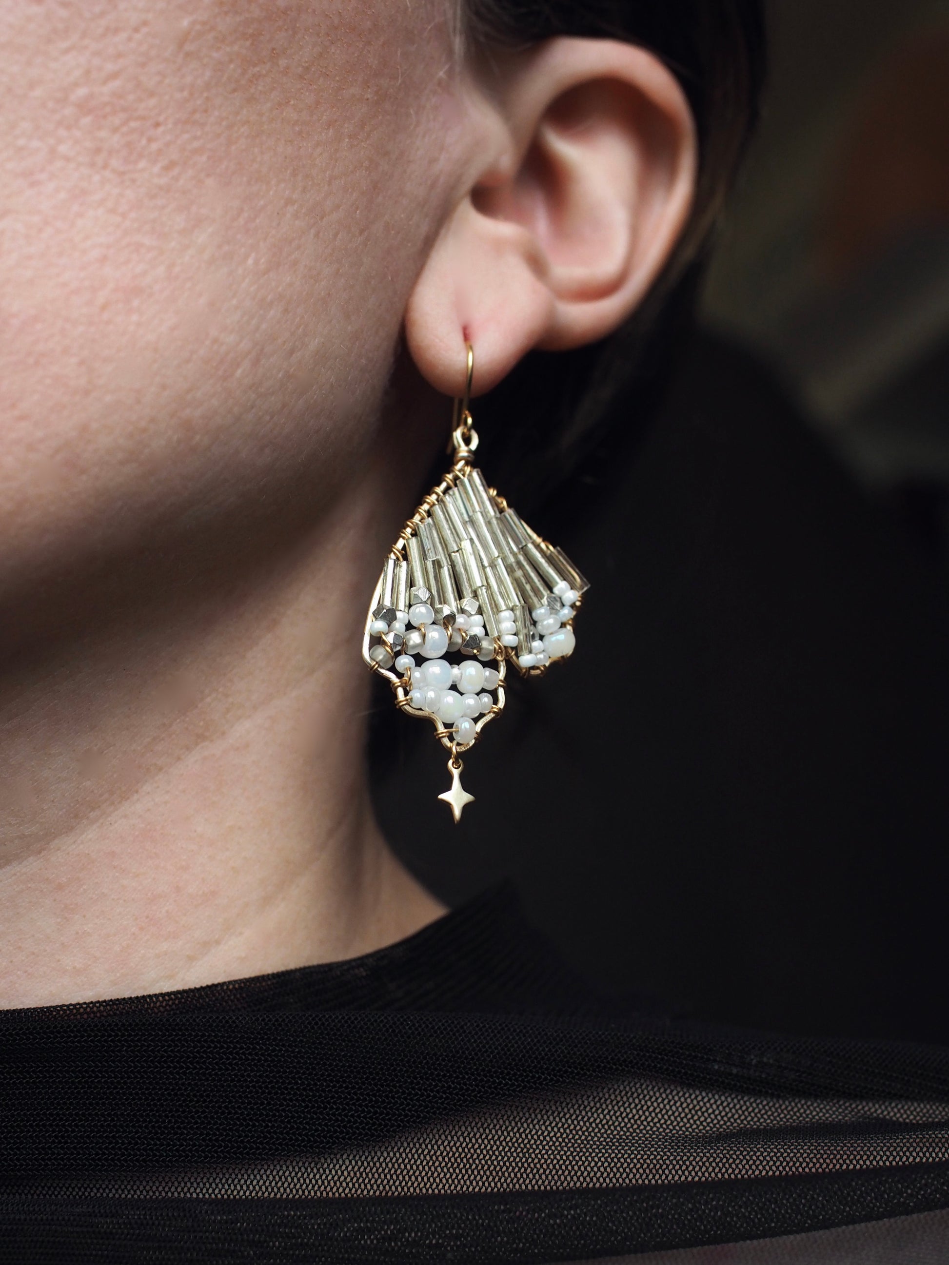 A close-up view of a person wearing a handmade celestial earring featuring a delicate wing-shaped design. The earring is crafted with shimmering metallic beads, opalescent accents, and a small dangling star charm, hanging elegantly from a gold hook.