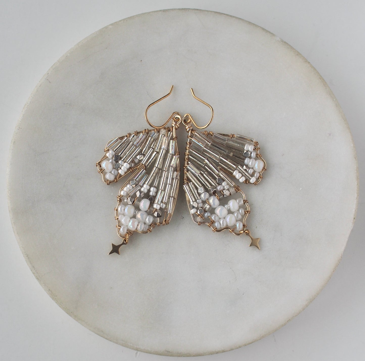 A close-up view of handmade celestial earring featuring a delicate wing-shaped design on a round marble dish positioned to look like a butterfly. Crafted with shimmering metallic beads, opalescent accents, and small dangling stars.