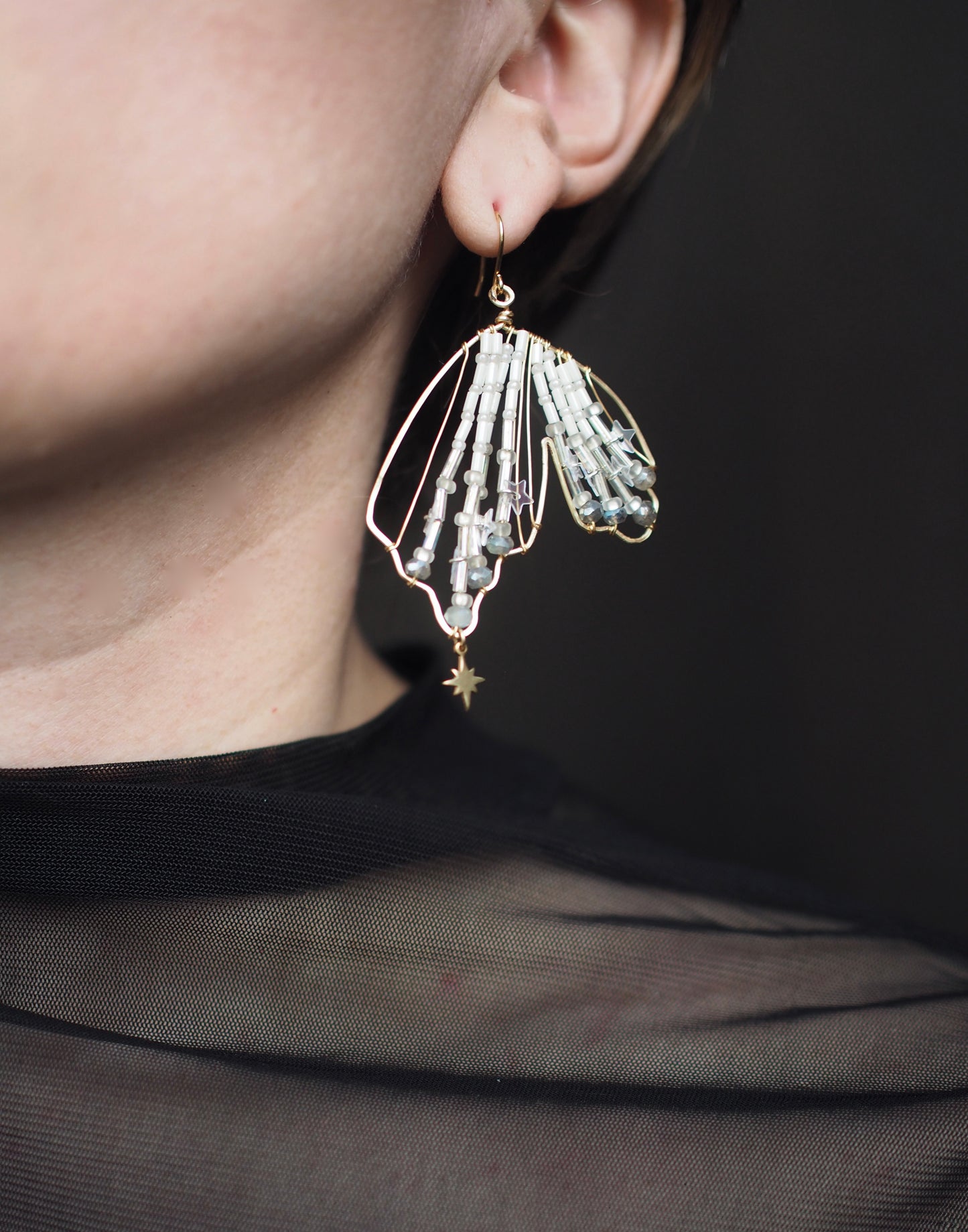 A close-up view of someone wearing a handmade celestial earring featuring a delicate wing-shaped design. The elegant earrings are crafted with shimmering metallic star sequins, opalescent beads, and small dangling stars.