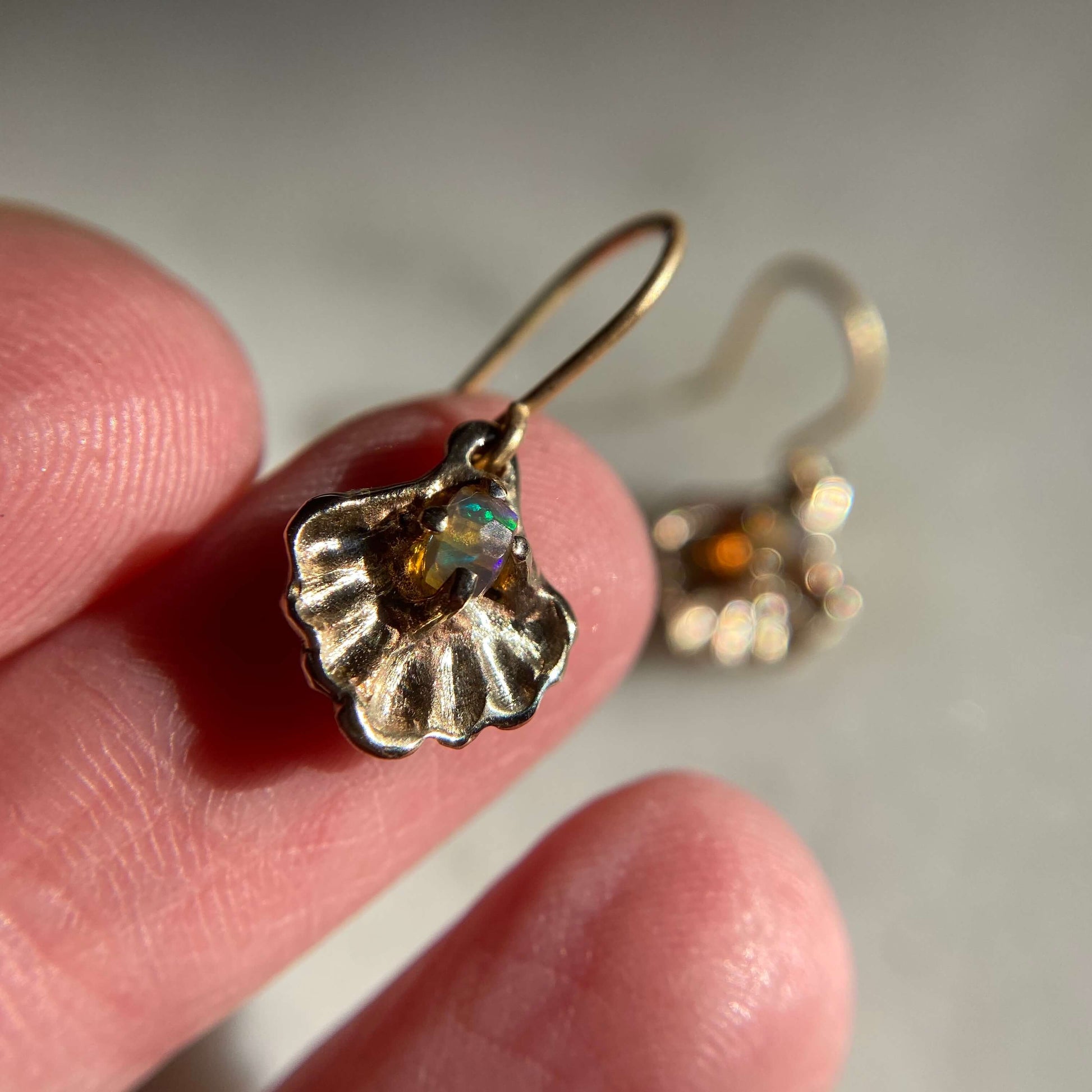 Oyster shell earrings containing a small opal "pearl", cast in gold tone bronze by Iron Oxide Designs, held on a fingertip to show scale