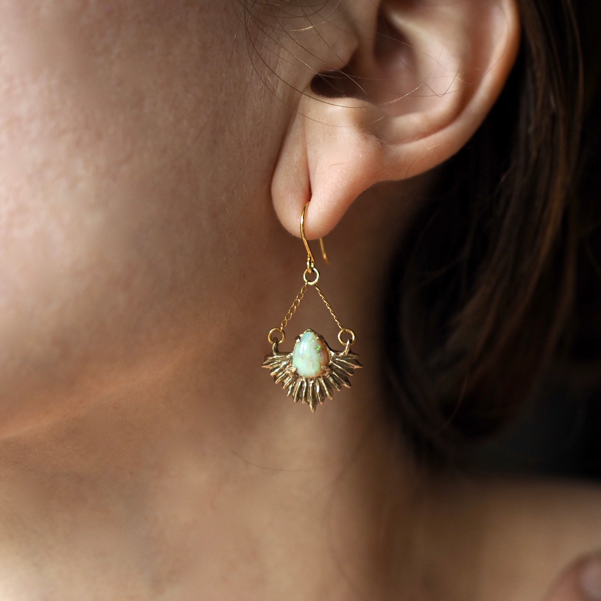 Gold Tone bronze earrings in the shape of beams of light set with teardrop shaped, lab grown opals on a model for scale